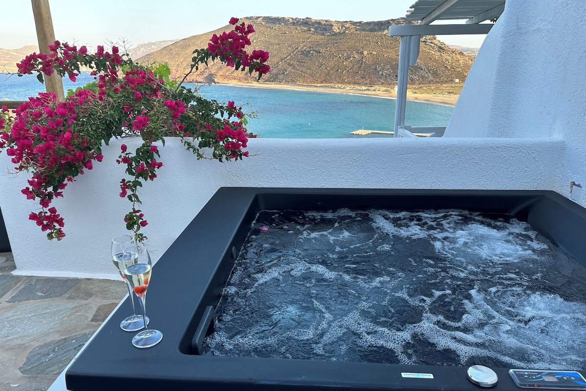 A hot tub with a view of the sea and mountains, a glass of champagne, and pink flowers.