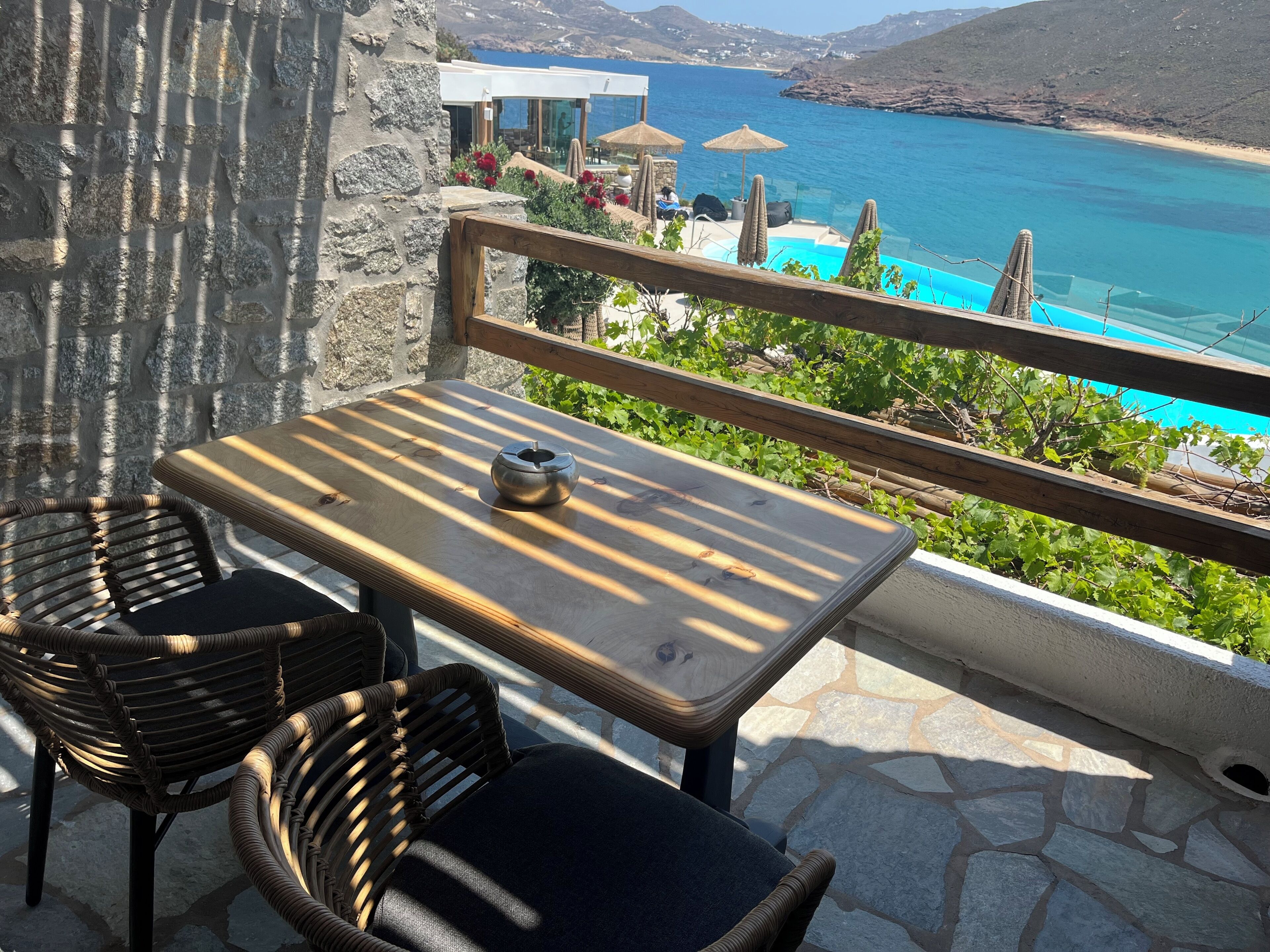 A terrace with a table and chairs, overlooking a coastal view with a pool and umbrellas.