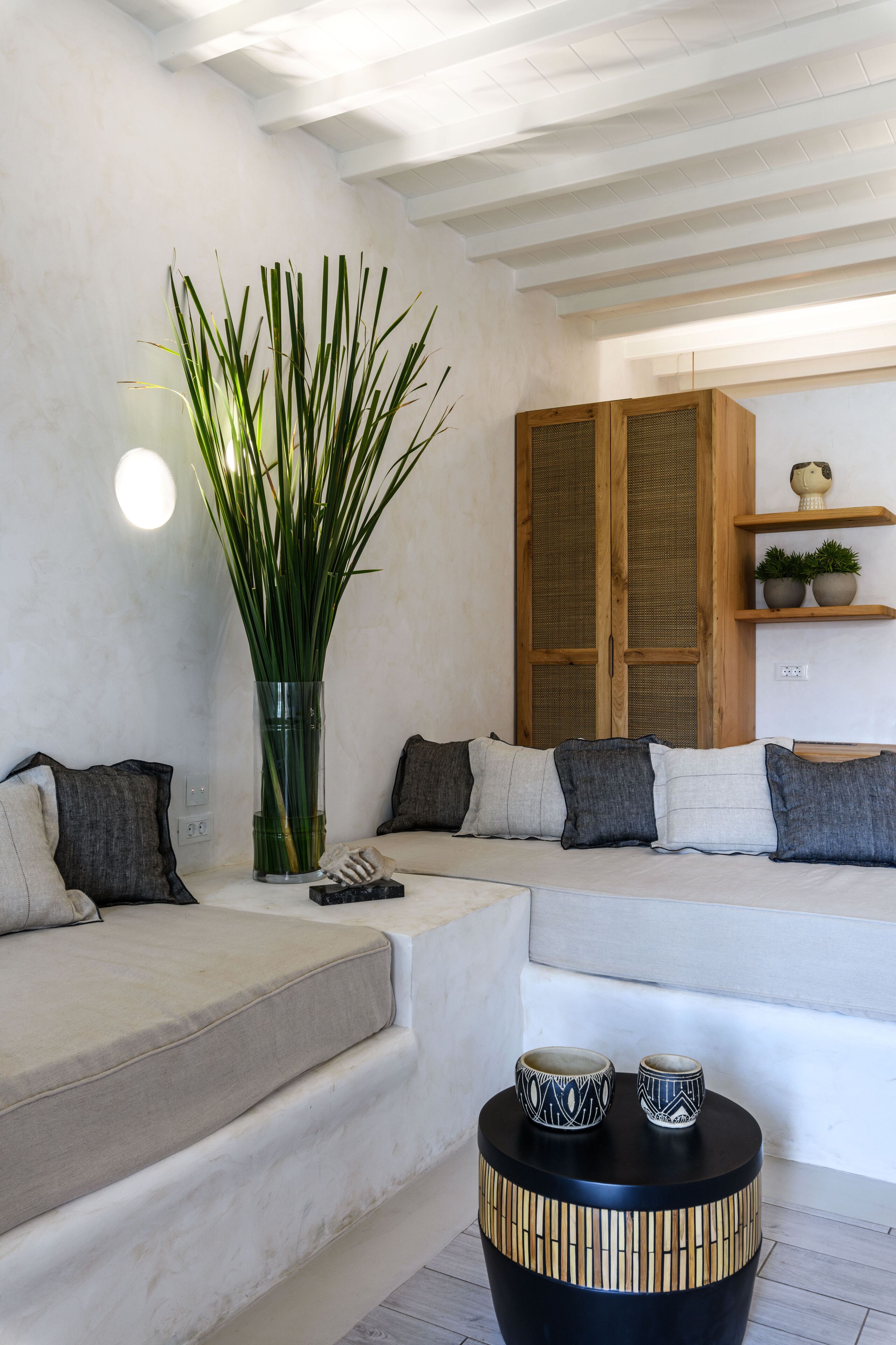 Ein modernes Wohnzimmer mit einer Couch, einer hohen, grünen Pflanze, einem Holzkabinett und einem kleinen Tisch mit dekorativen Schalen.