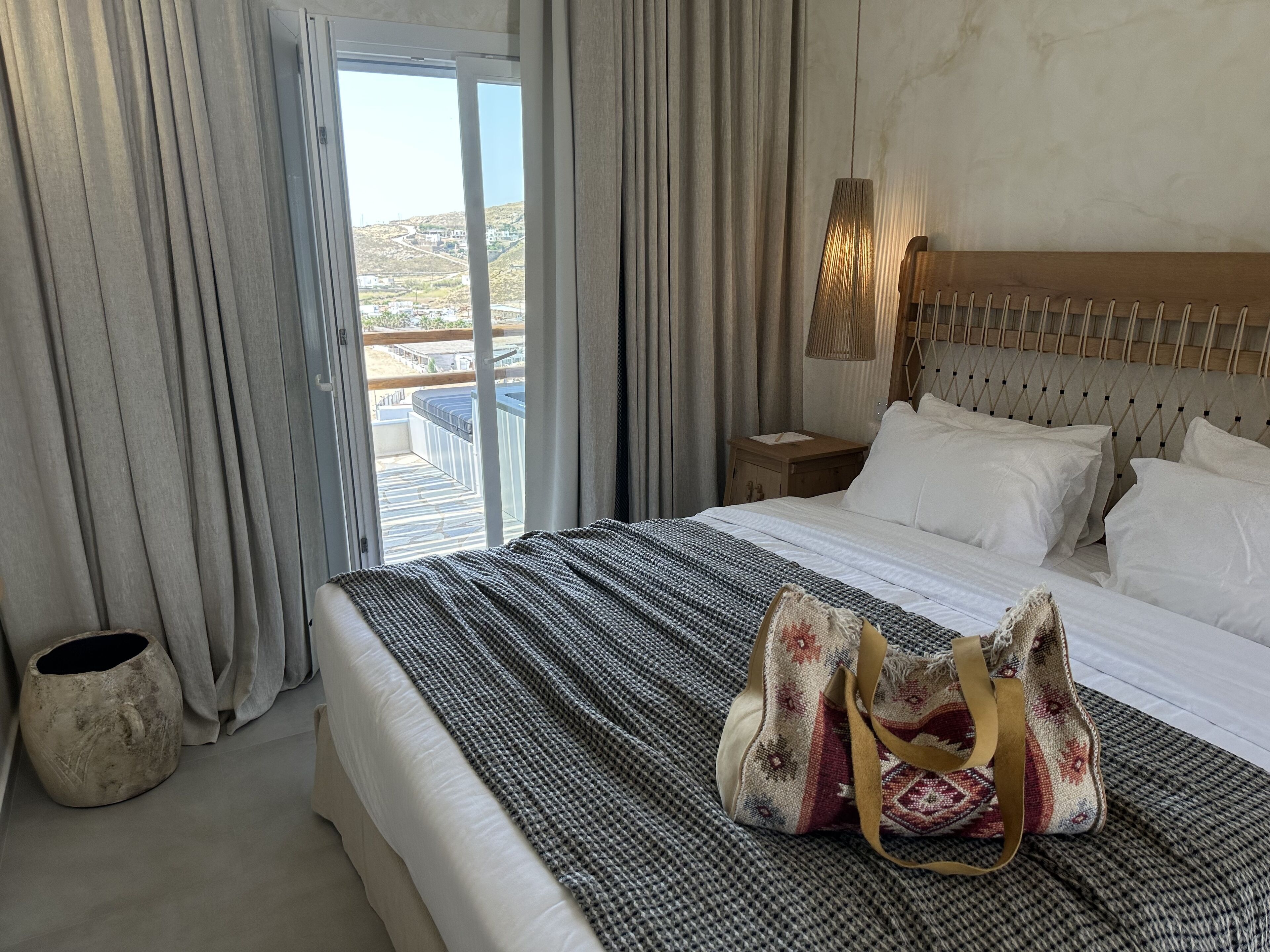 Habitación de hotel con cama, mesita de noche, lámpara, vista al exterior y un amplio ventanal.