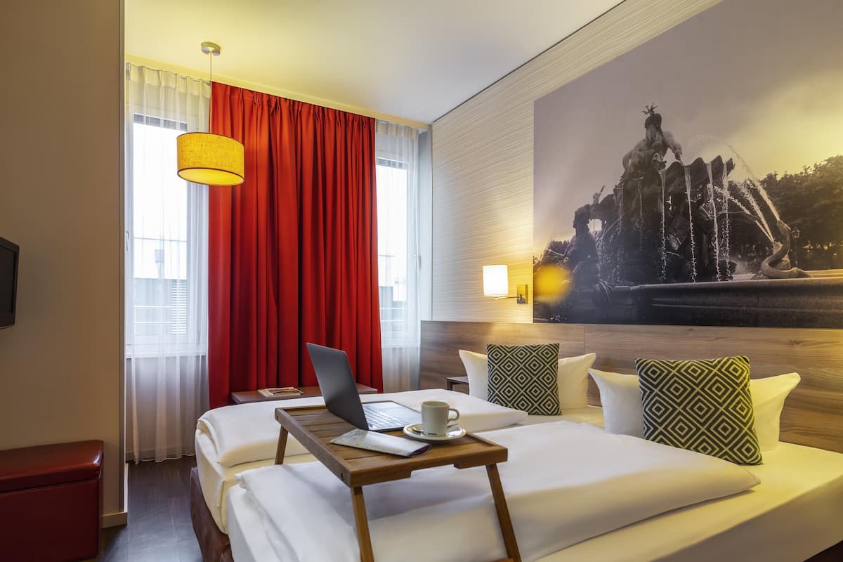 A hotel room with a bed, a laptop, a tray table, a red curtain, a wall-mounted lamp, and a large painting of a fountain.