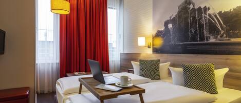 A hotel room with a bed, a laptop, a tray table, a red curtain, a wall-mounted lamp, and a large painting of a fountain.