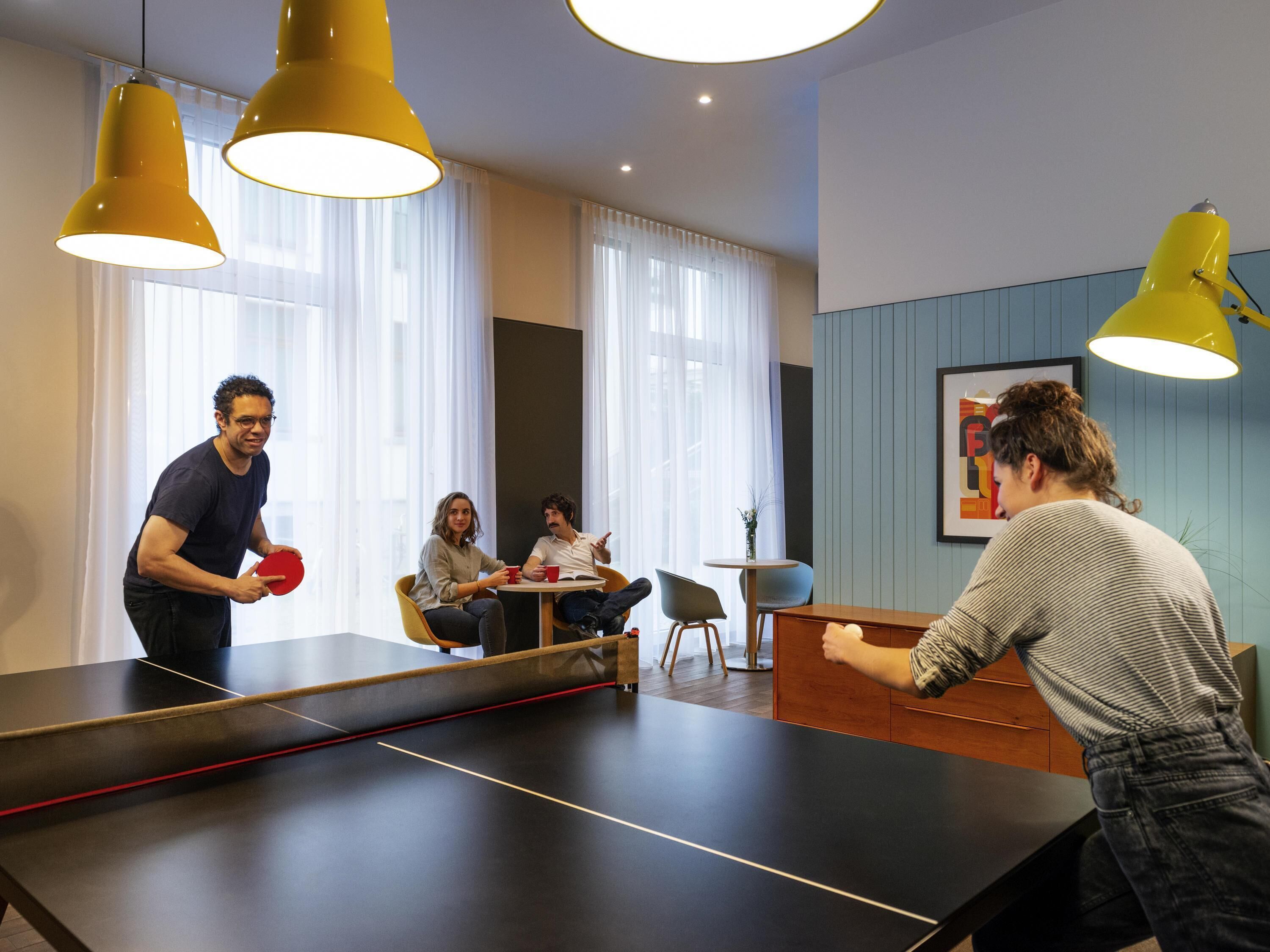 A ping pong table in a modern room with large windows, yellow pendant lights, and a small table with two people seated.