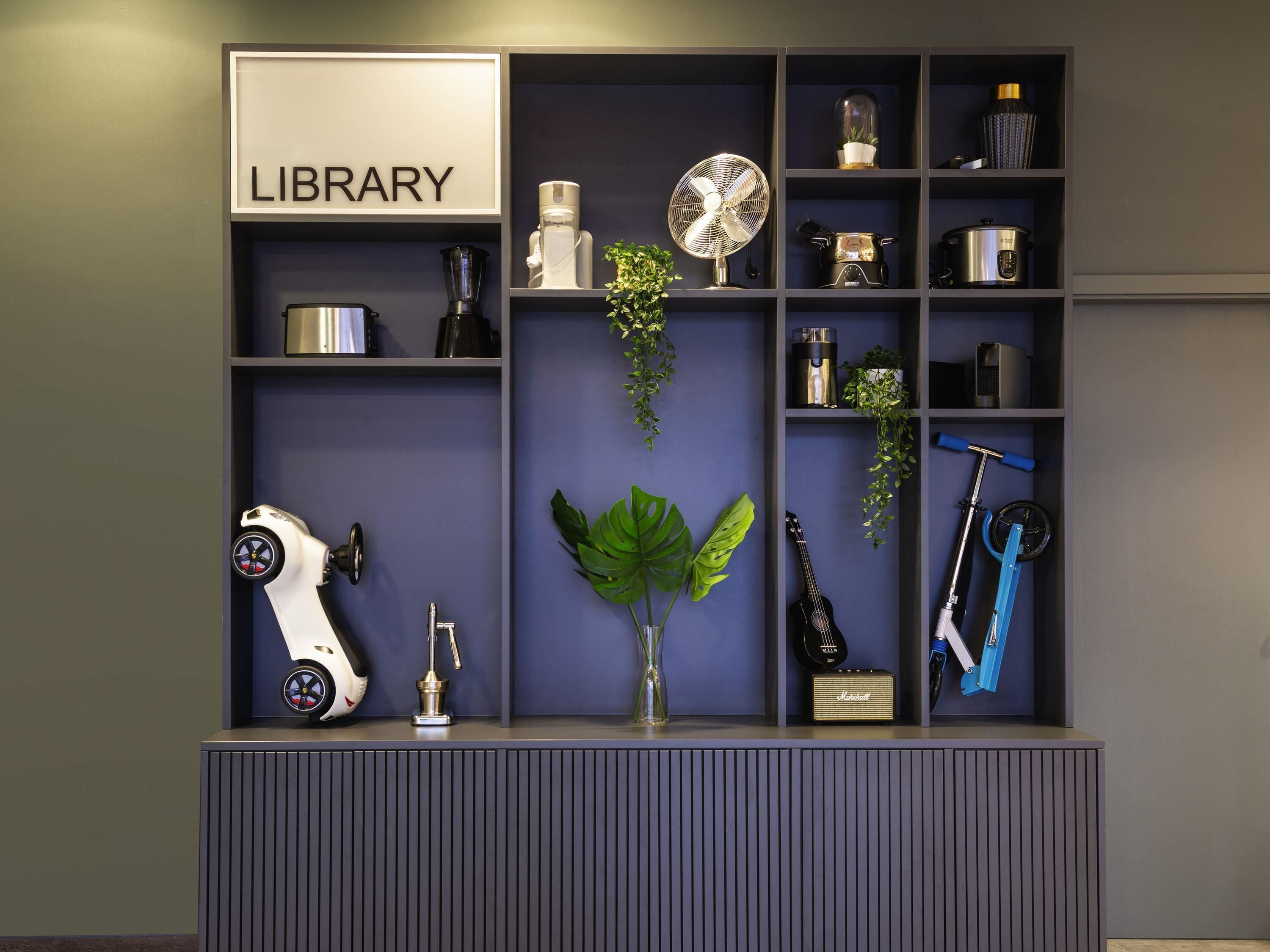 Una habitación con un letrero que dice 'LIBRARY', un scooter eléctrico blanco, una guitarra, un amplificador Marshall y un scooter azul, además de una planta.