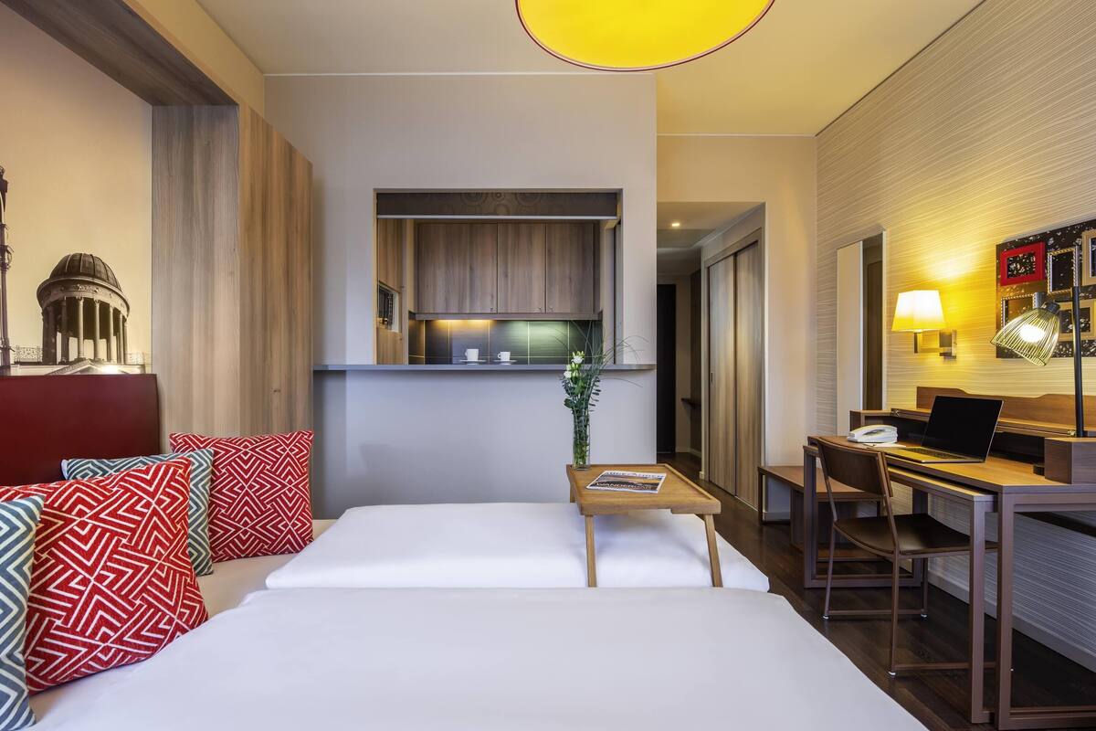 A hotel room with a bed, a desk, a chair, a lamp, a small table, and a view of the kitchen area.