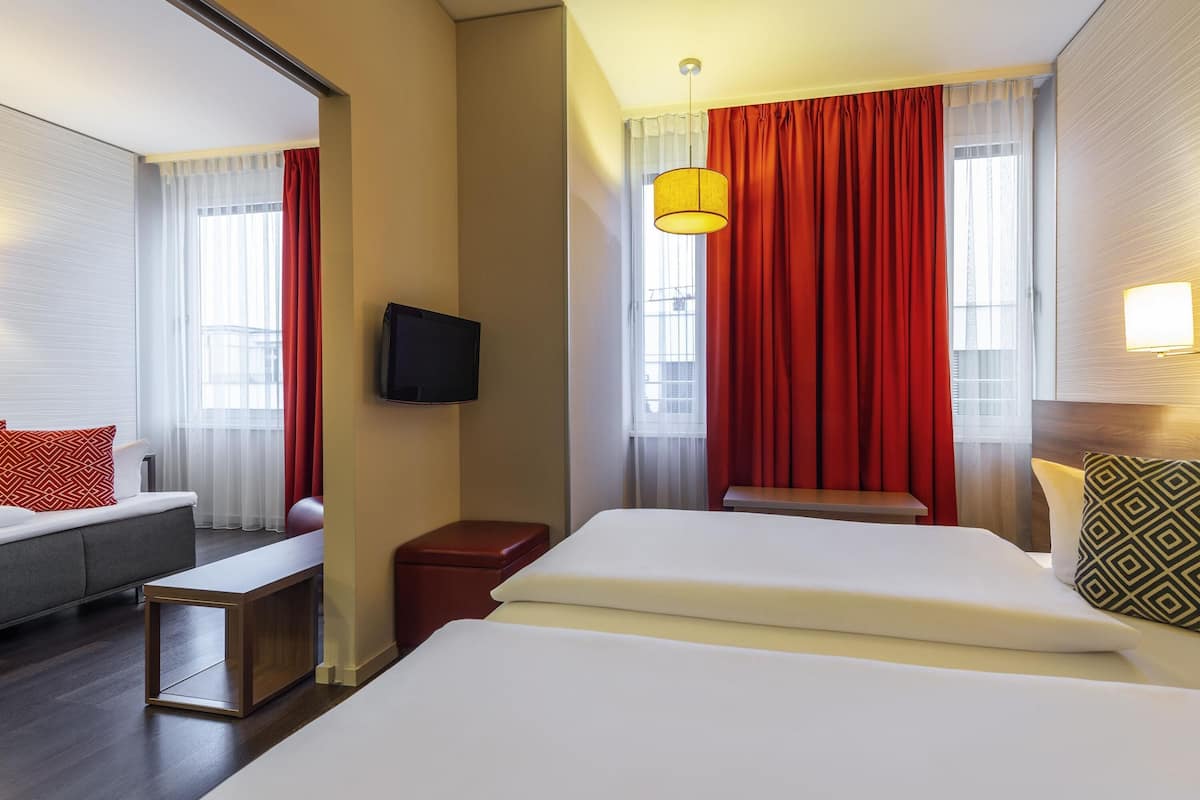 Una camera d'albergo con un letto, una poltrona, una TV, un tavolino e una finestra con tende.