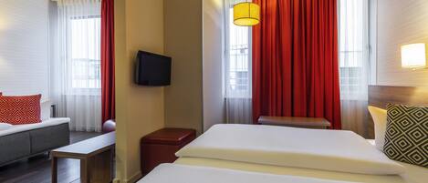 A hotel room with a bed, a sofa, a TV, a small table, and a window with curtains.