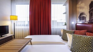 Ein Hotelzimmer mit zwei Betten, einem Fernseher, einer Lampe und Blick auf ein Gebäude durch das Fenster.