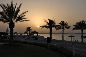 A beach scene with palm trees, a gazebo, and a distant view of a bridge. - Al Marfa Hotel (Al Mirfa)