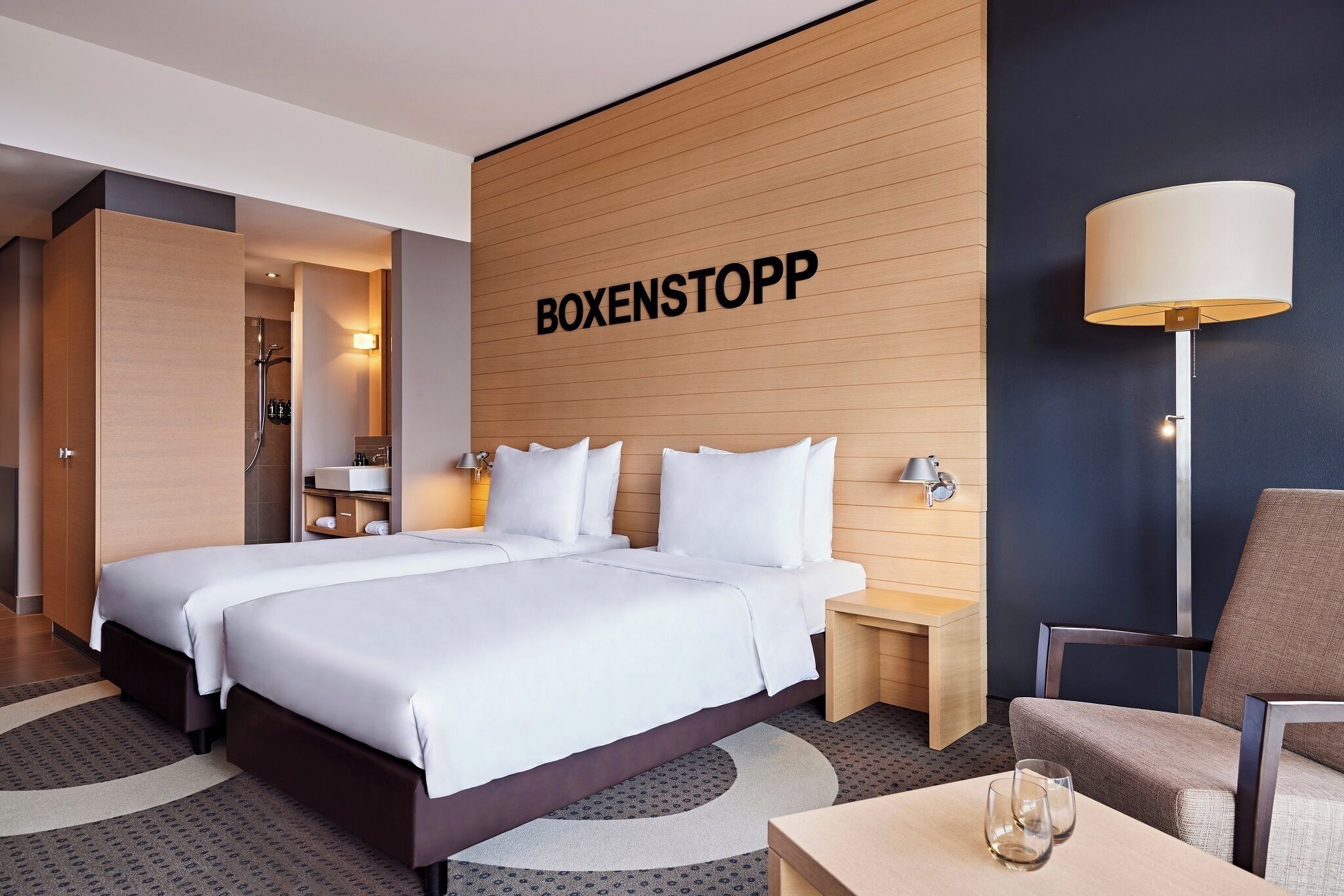 A hotel room with two beds, a wooden accent wall, a lamp, and a chair.