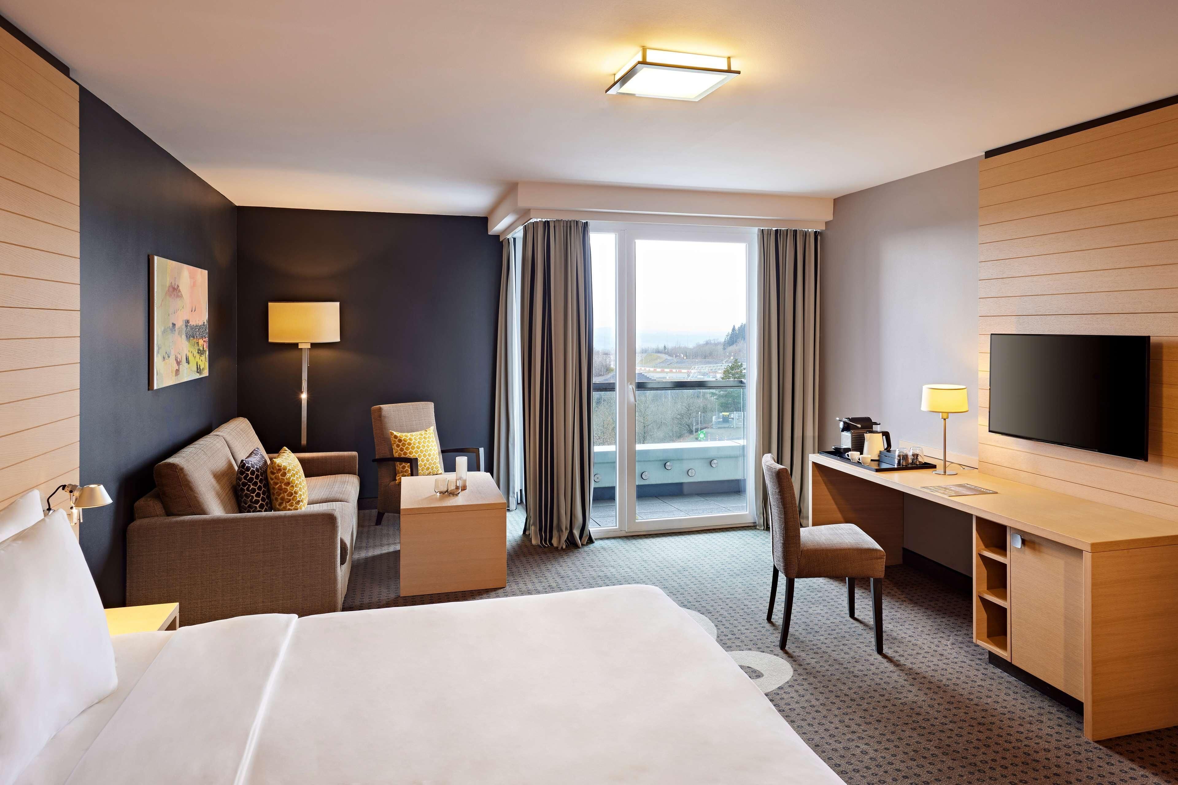 Une chambre d’hôtel avec un grand lit, un bureau avec une télévision, un canapé, un fauteuil et une vue sur la ville par la fenêtre.
