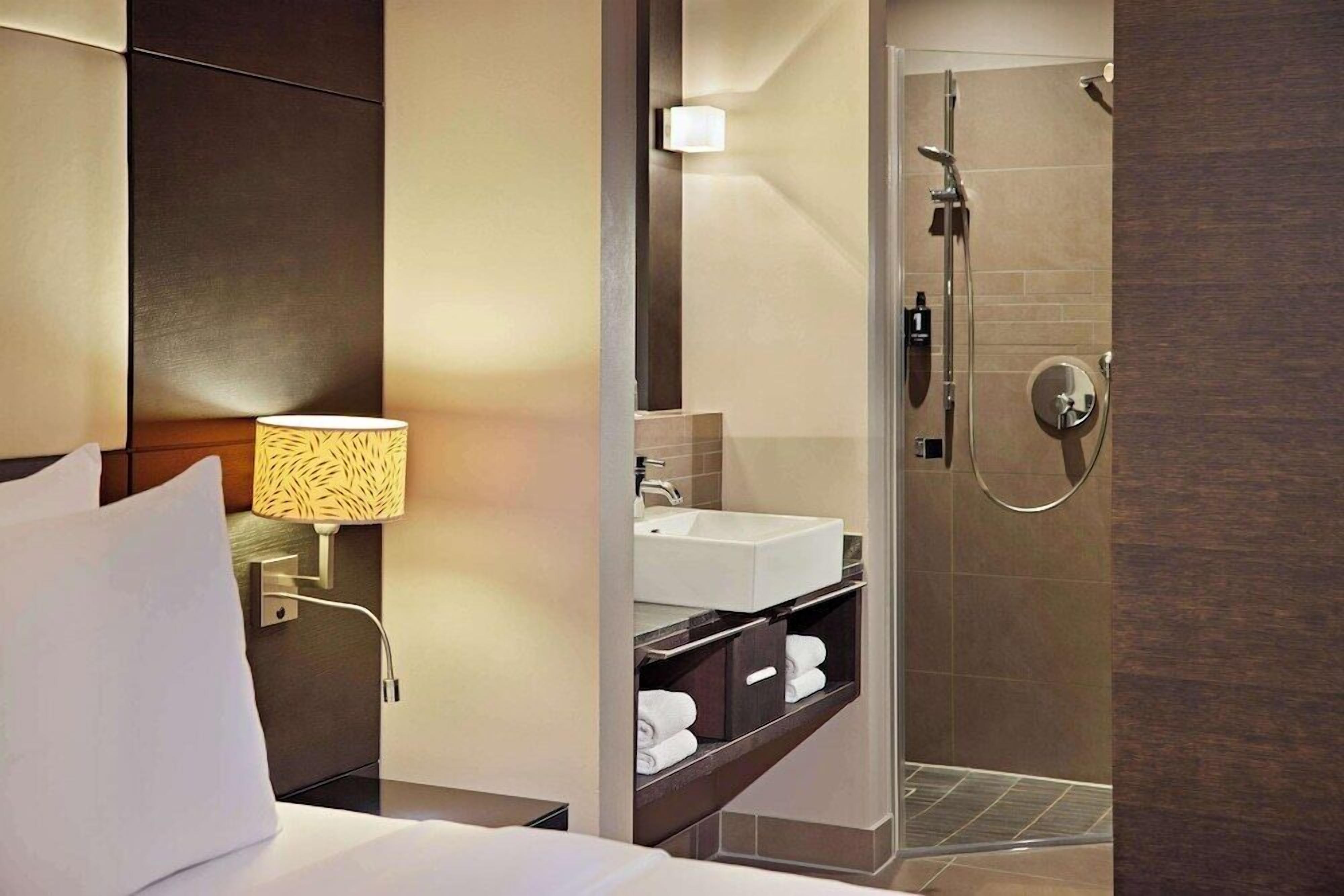 A hotel room with a bed, a bedside lamp, a bathroom with a sink, a shower, and a mirror.