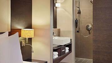 A hotel room with a bed, a bedside lamp, a bathroom with a sink, a shower, and a mirror.