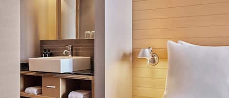 Una habitación de hotel moderna con una cama grande, un tocador empotrado con espejo, un lavabo y una mesita de noche con lámpara.