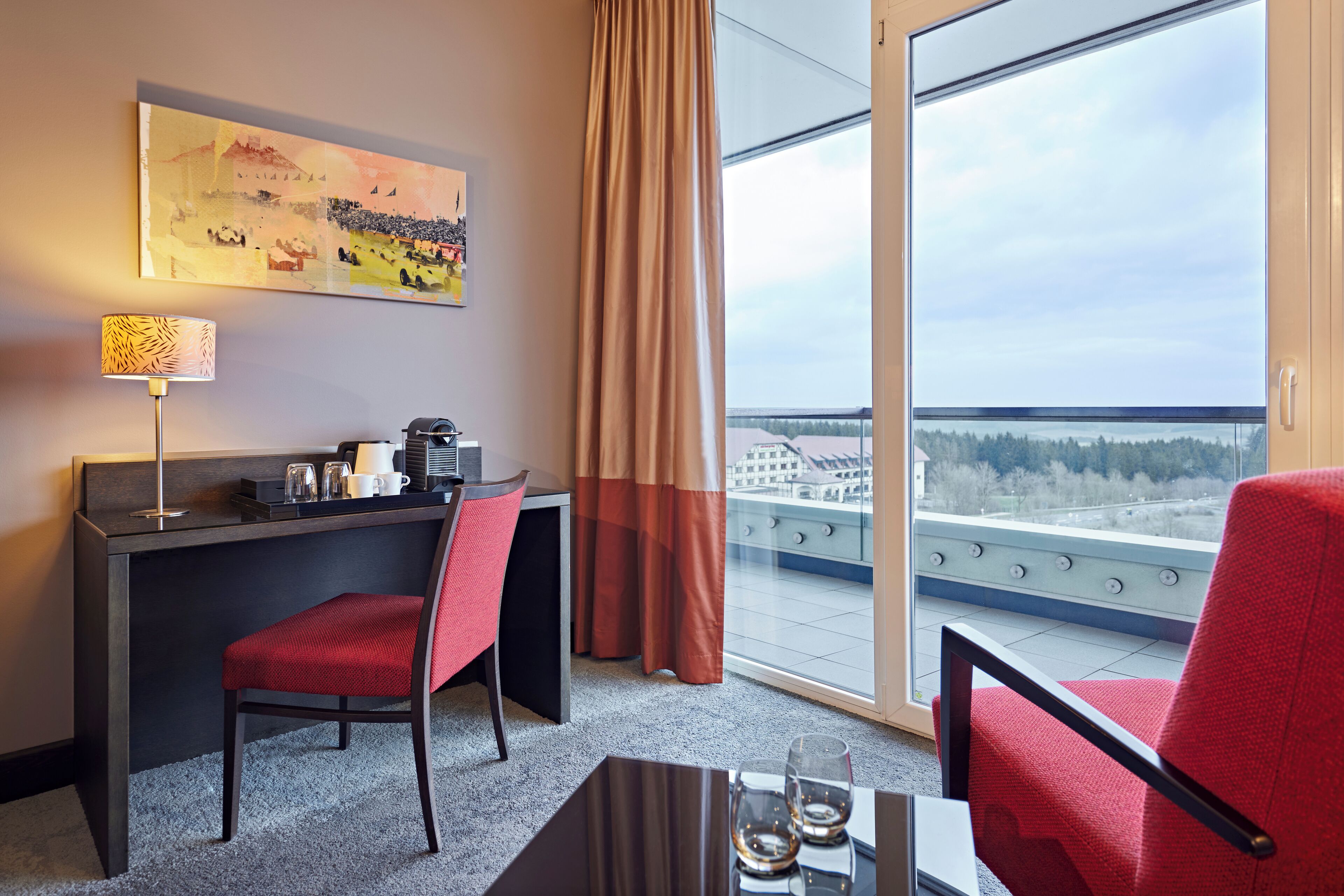 Quarto de hotel com escrivaninha, duas cadeiras vermelhas, máquina de café, luminária e janela grande com vista para a paisagem urbana e árvores.