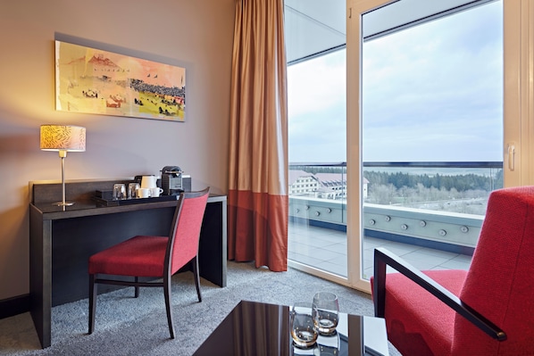 Una habitación de hotel con escritorio, dos sillas rojas, cafetera, lámpara y ventana grande con vista a la ciudad y árboles.