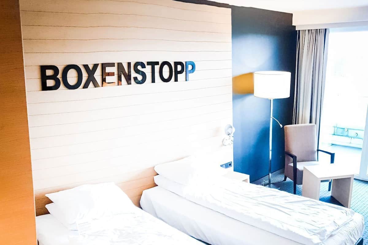 A hotel room with two beds, a headboard with the name 'BOXENSTOPP', a desk, a chair, and a lamp.