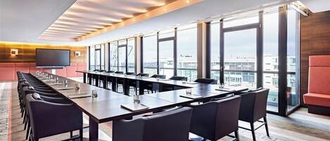 A conference room with a long table, multiple chairs, a flat-screen TV, and large windows offering a city view.