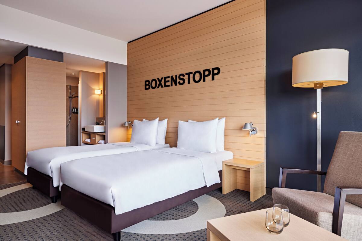 A hotel room with two beds, a wooden accent wall, a lamp, and a chair.