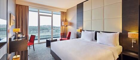 Habitación de hotel con una cama grande, un escritorio con silla, un ventanal con vistas y una mesita de noche con lámpara.