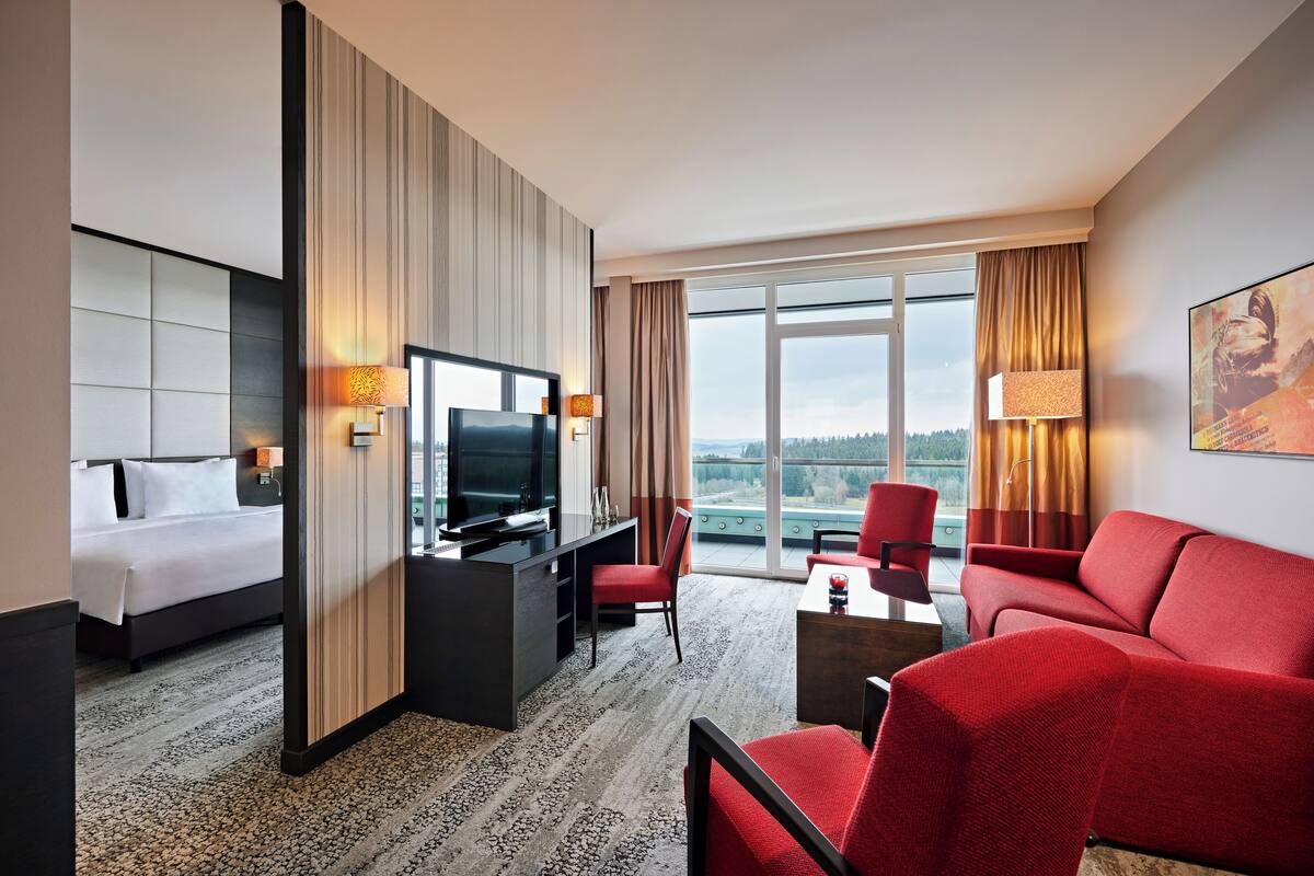 A hotel room with a bed, a desk with a TV, a red sofa, and a large window with a view.