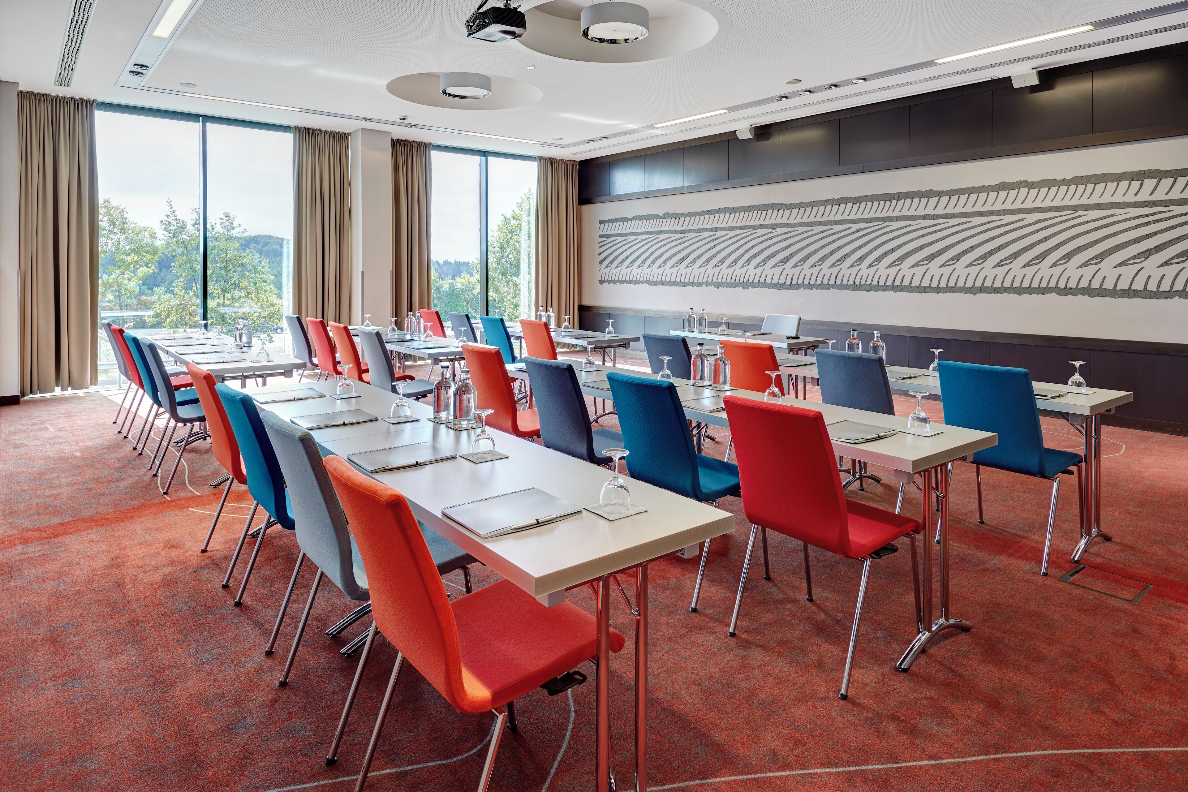 Un salón de conferencias con varias mesas y sillas, ventanas amplias y un diseño moderno en la pared.