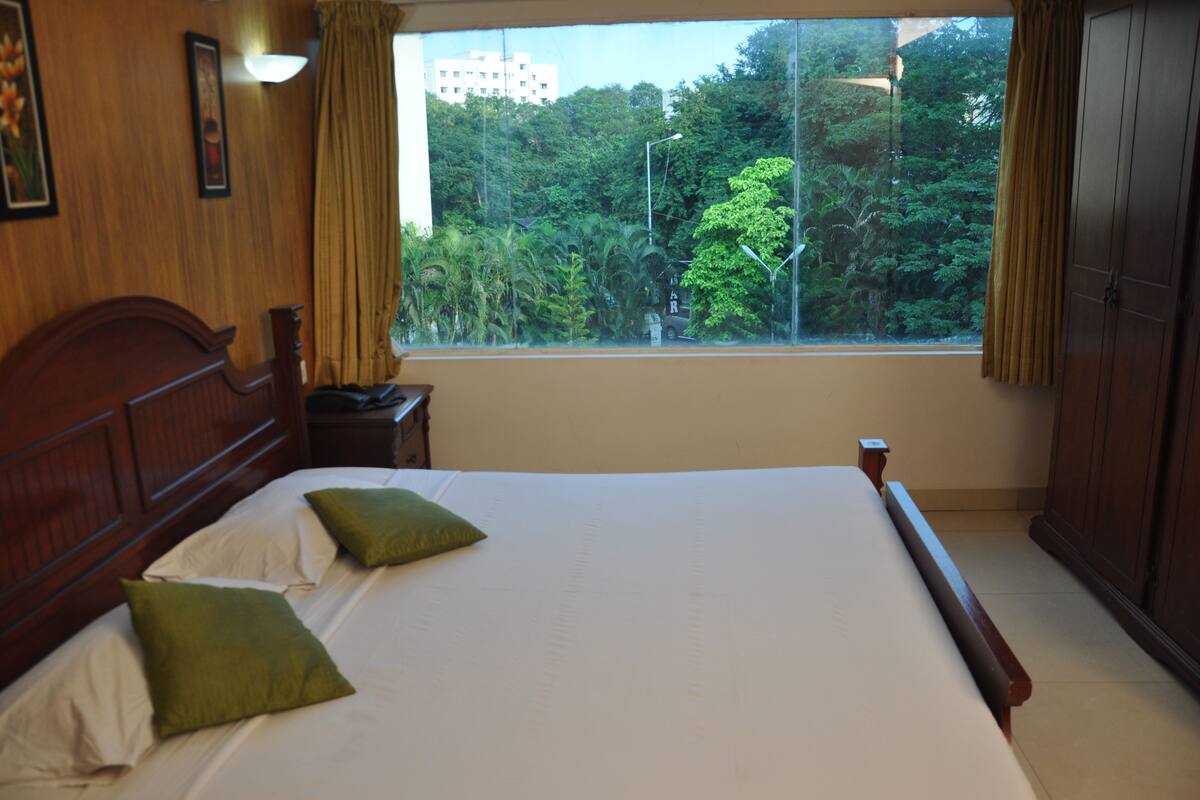 Een hotelkamer met een groot bed, een houten hoofdbord, twee groene kussens, een nachtkastje, een raam met gordijnen en uitzicht op bomen en gebouwen.