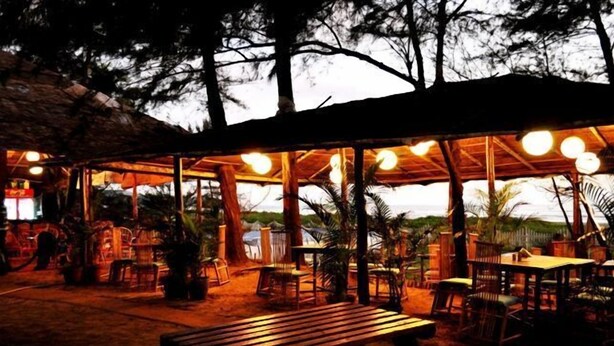 An outdoor patio area with wooden tables and chairs, surrounded by trees and a thatched roof structure. - Montego Bay Beach Village (Morjim)