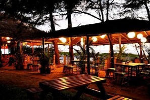An outdoor patio area with wooden tables and chairs, surrounded by trees and a thatched roof structure. - Montego Bay Beach Village (Morjim)