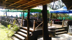 An outdoor seating area with wooden benches and tables, surrounded by green canopies and a bamboo structure.