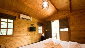 A wooden cabin room with a bed, a mounted television, and a window with a view of greenery. - Montego Bay Beach Village (Morjim)