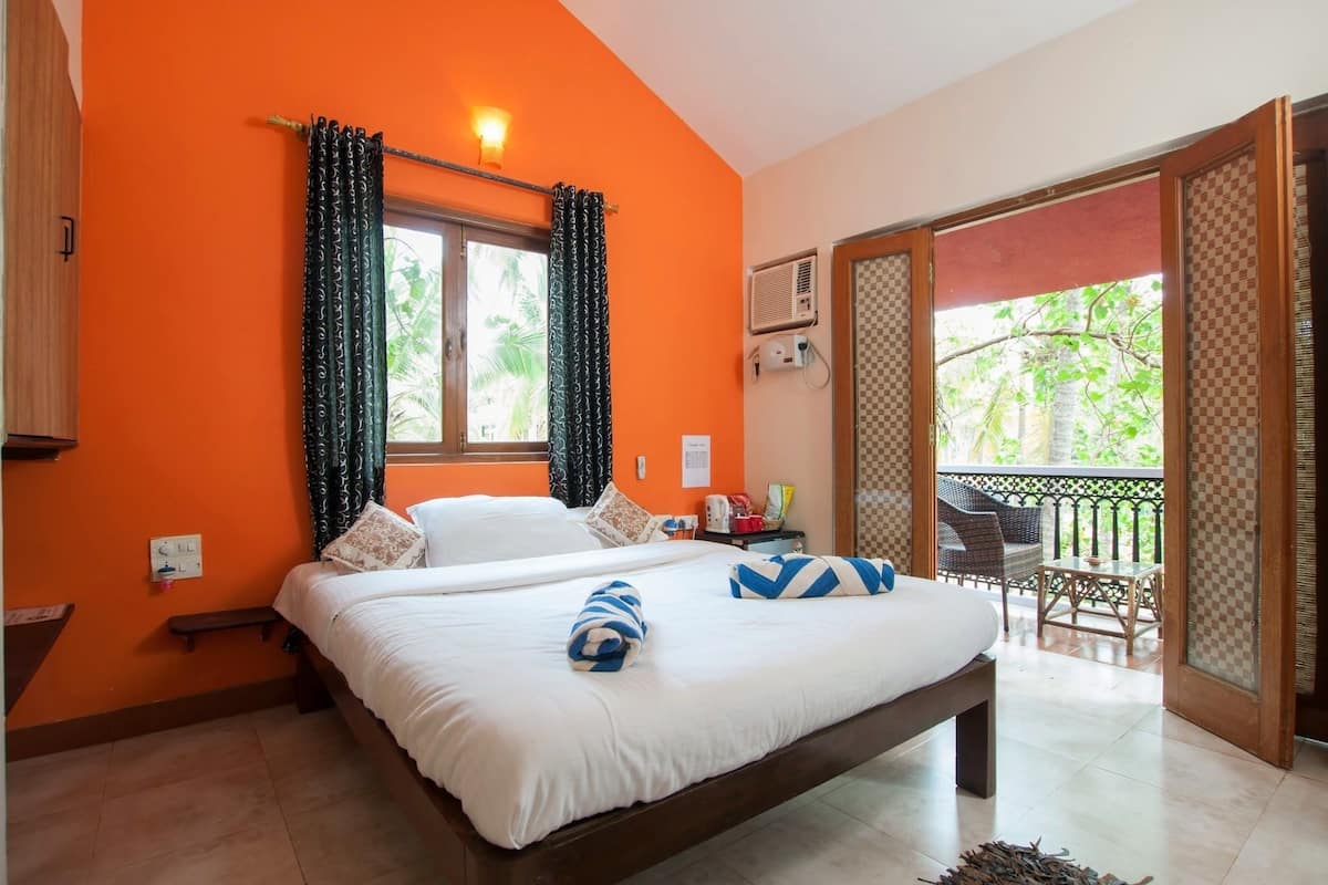 Quarto com parede laranja, cama com roupa de cama branca, um banco, janela com cortinas estampadas e varanda com móveis de exterior.