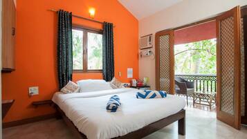 Un dormitorio con pared naranja, una cama con ropa blanca, un banco, una ventana con cortinas estampadas y un balcón con muebles de exterior.