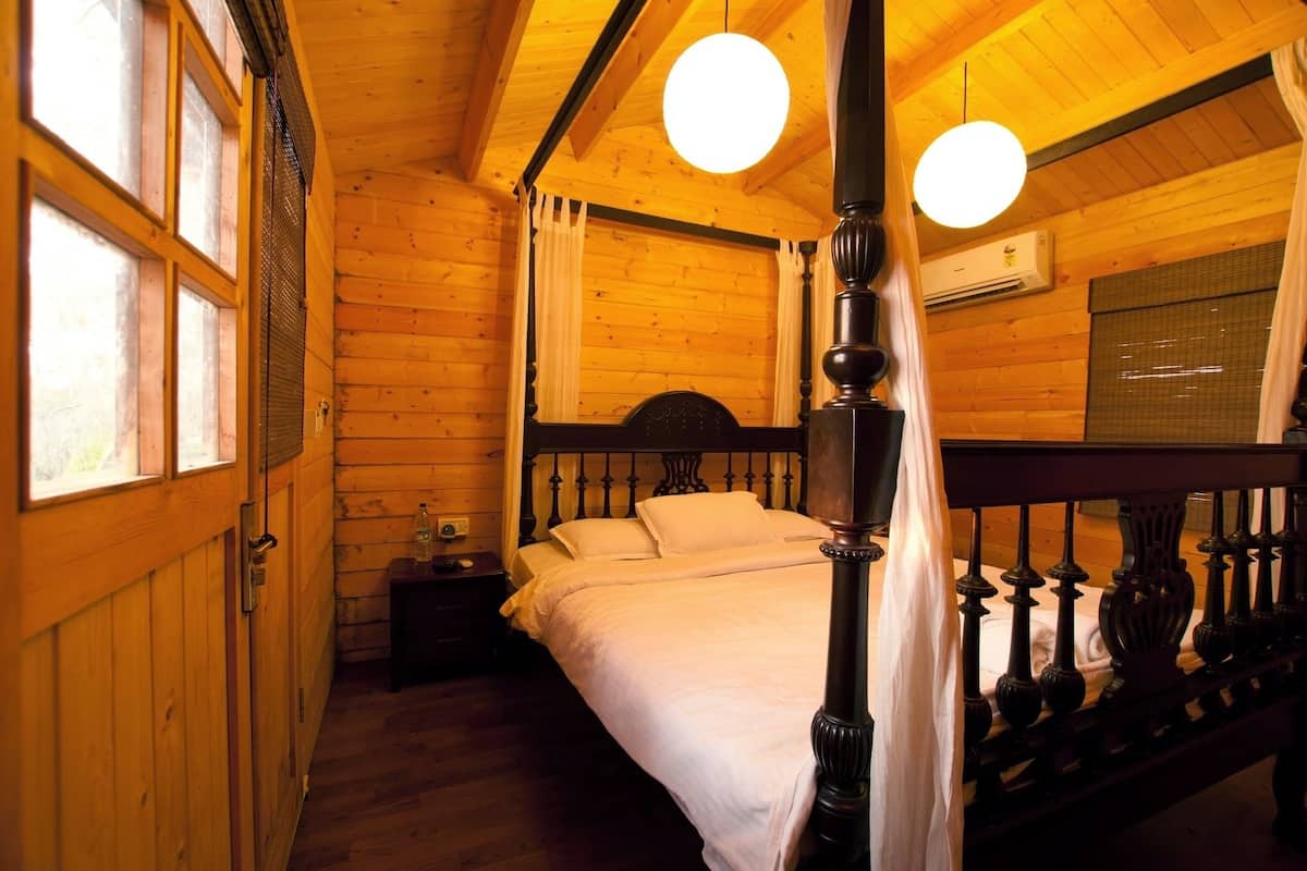 Uma cama de madeira com dossel, roupa de cama branca, uma mesinha de cabeceira com abajur e uma janela com cortinas.