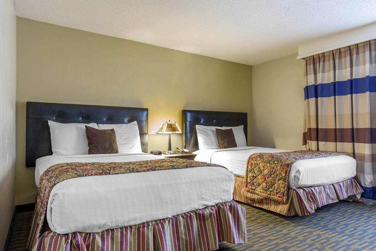 A hotel room with two beds, a nightstand, a lamp, and a window with curtains.