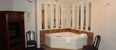 A hotel room with a large bathtub, a wooden cabinet, two chairs, a mirror wall, and a ceiling-mounted light fixture.