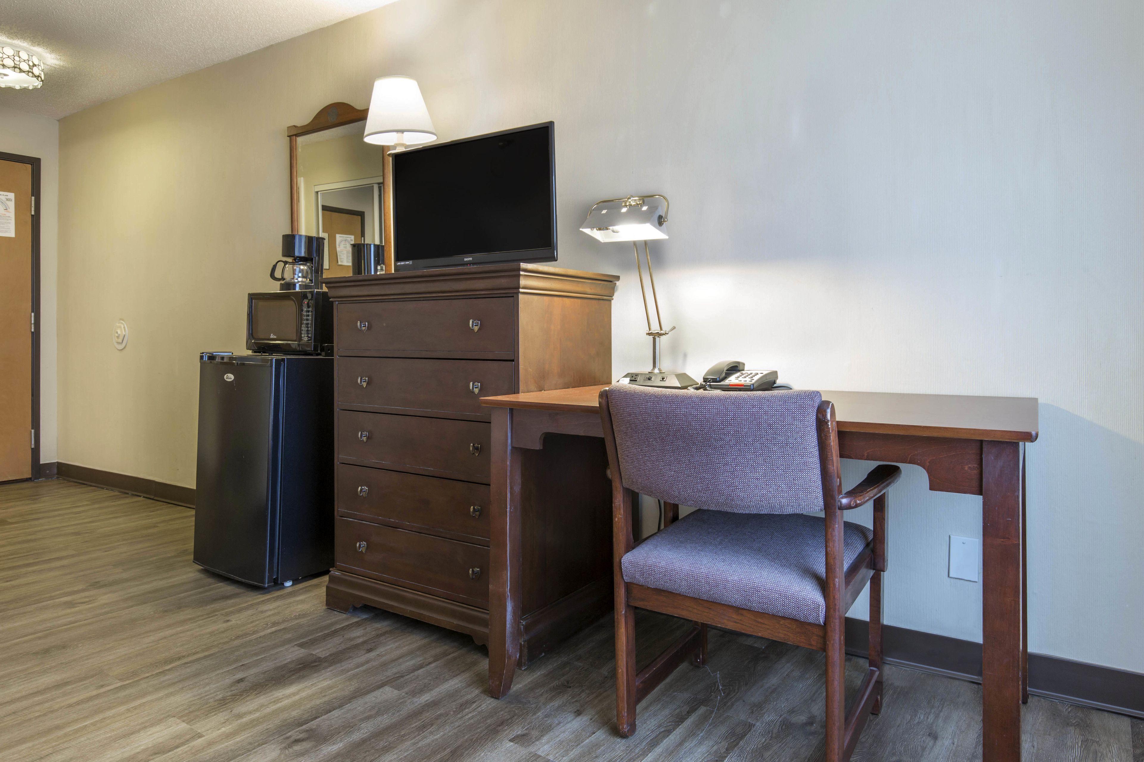 A hotel room with a wooden desk, a chair, a dresser, a microwave, a television, and a lamp.