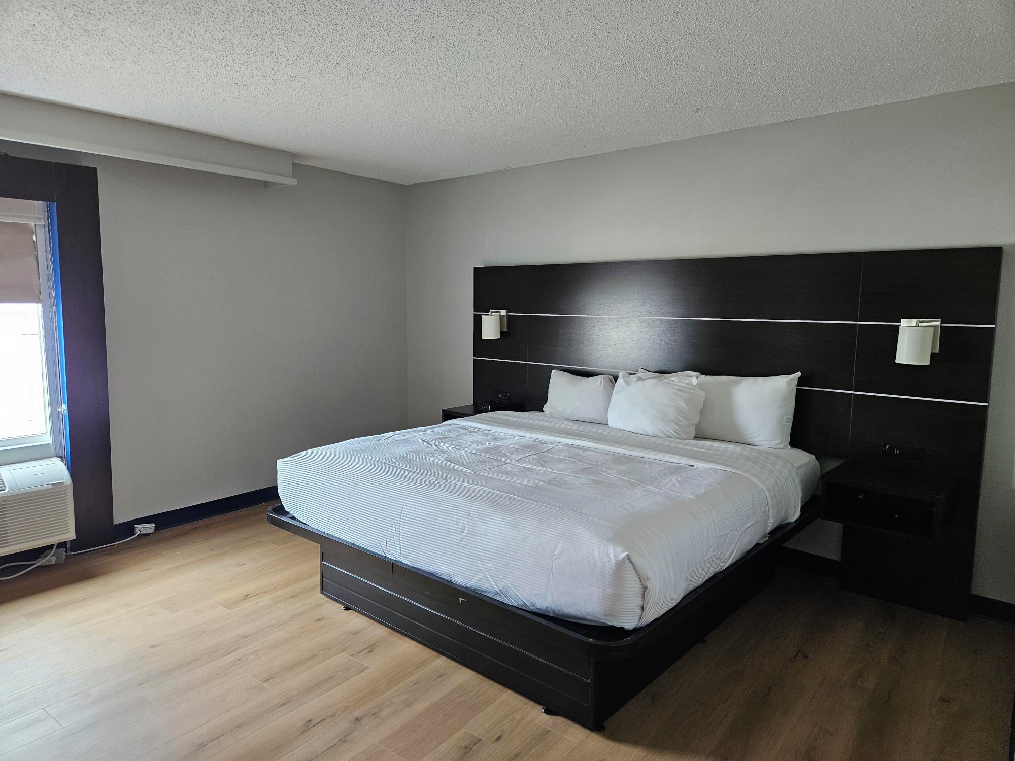 Un dormitorio moderno con una cama grande, cabecero oscuro, lámparas de noche y una ventana con persianas.