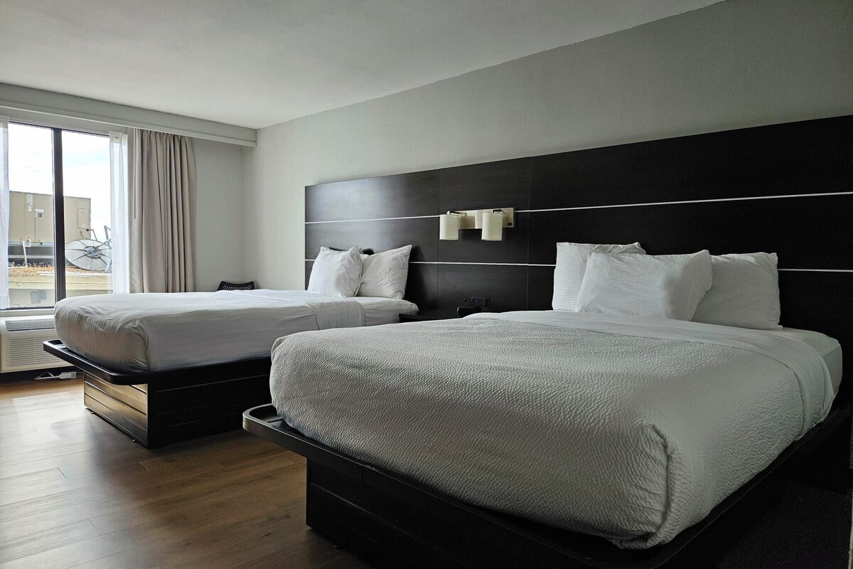 A hotel room with two beds, a large window with curtains, and a view of the outside.