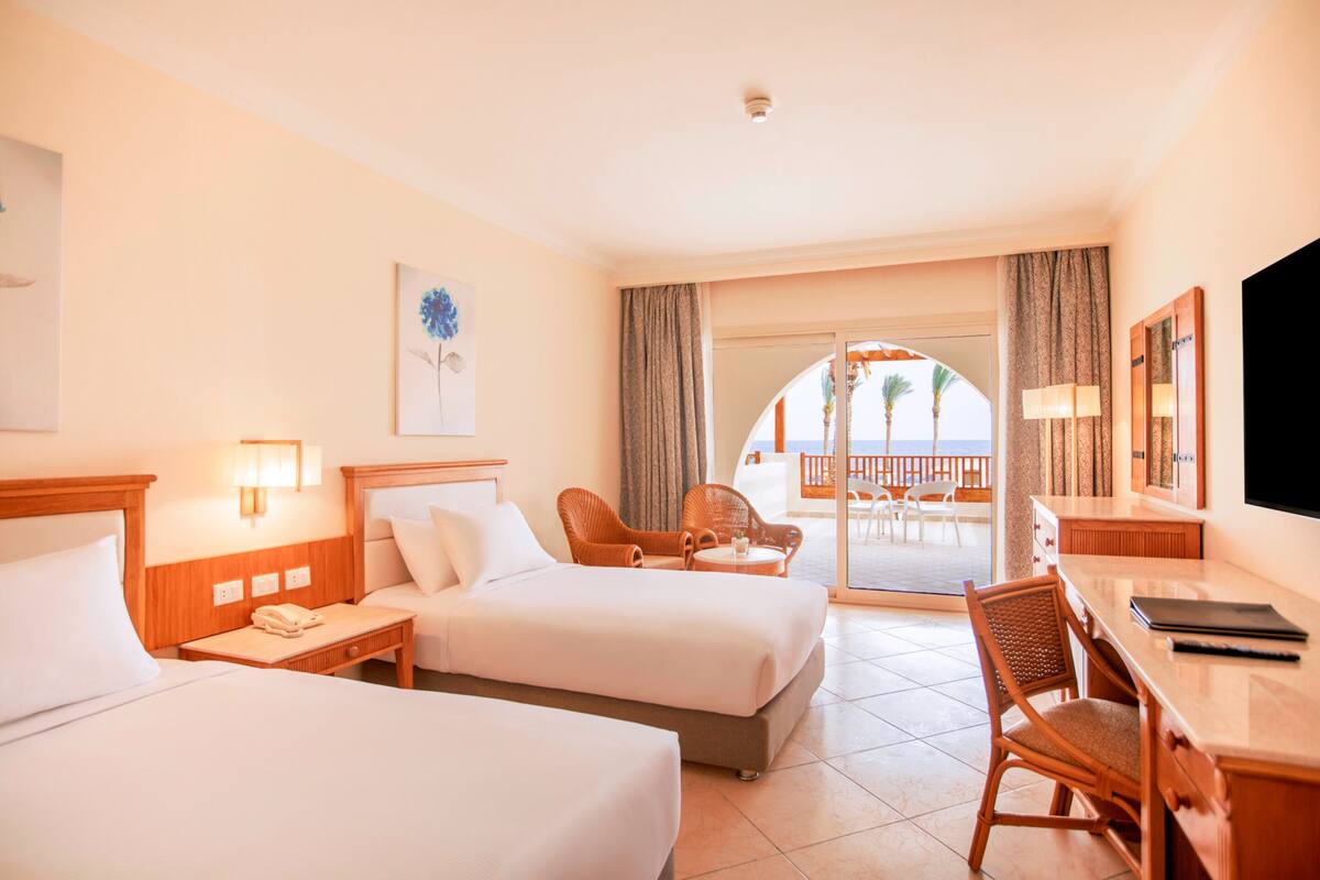 A hotel room with two beds, a desk, a chair, a TV, and a balcony with a view.