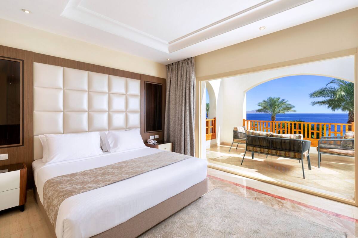 A hotel room with a large bed, a TV, a desk, and a balcony with a view of the sea.