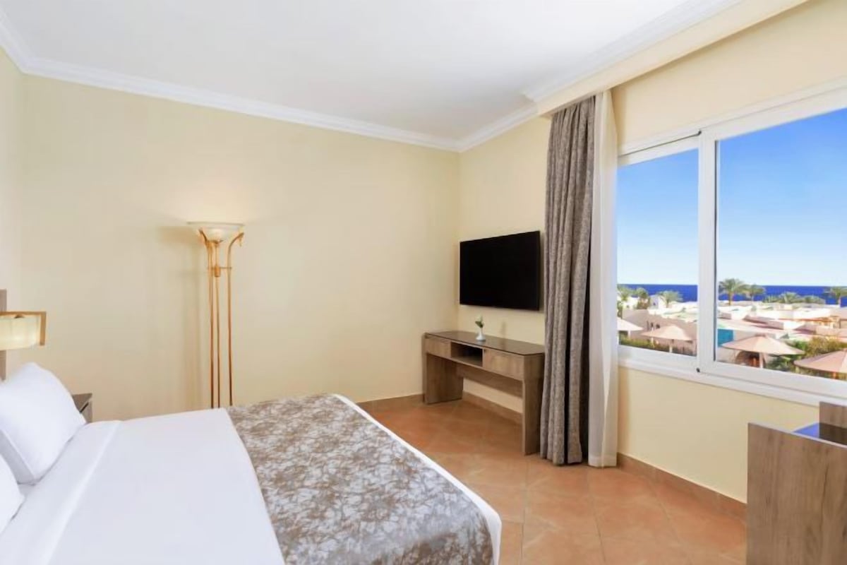 A hotel room with a bed, a desk with a flat-screen TV, a lamp, and a window with a view of the city.