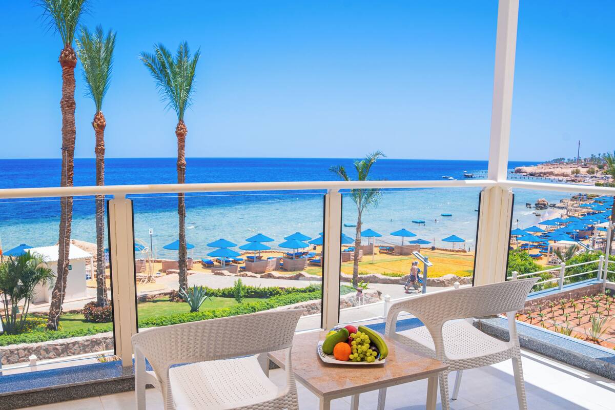 A balcony with two wicker chairs and a table with a fruit bowl. The view includes a beach with umbrellas and palm trees.