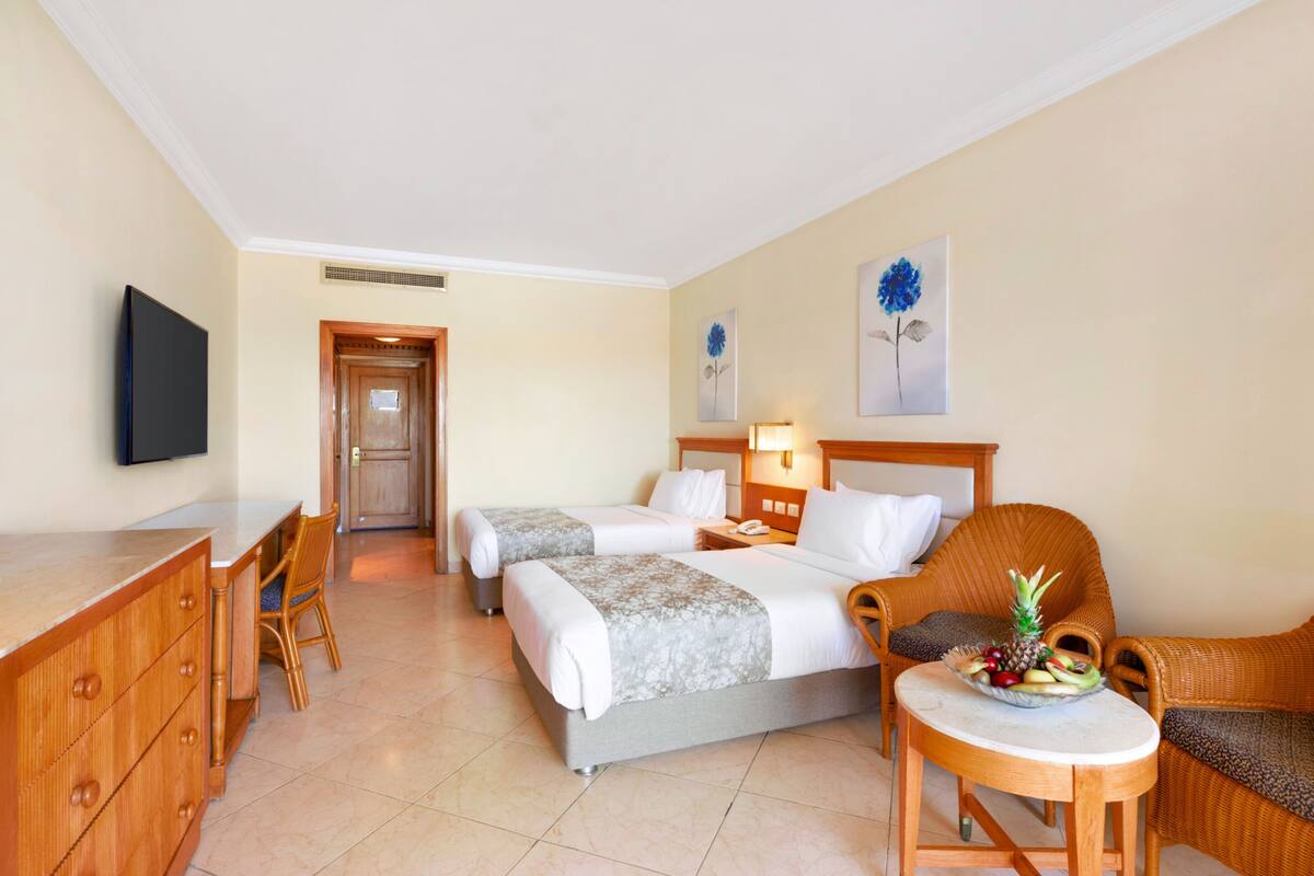 A hotel room with two beds, a desk, a chair, a small table with fruit, and artwork on the walls.