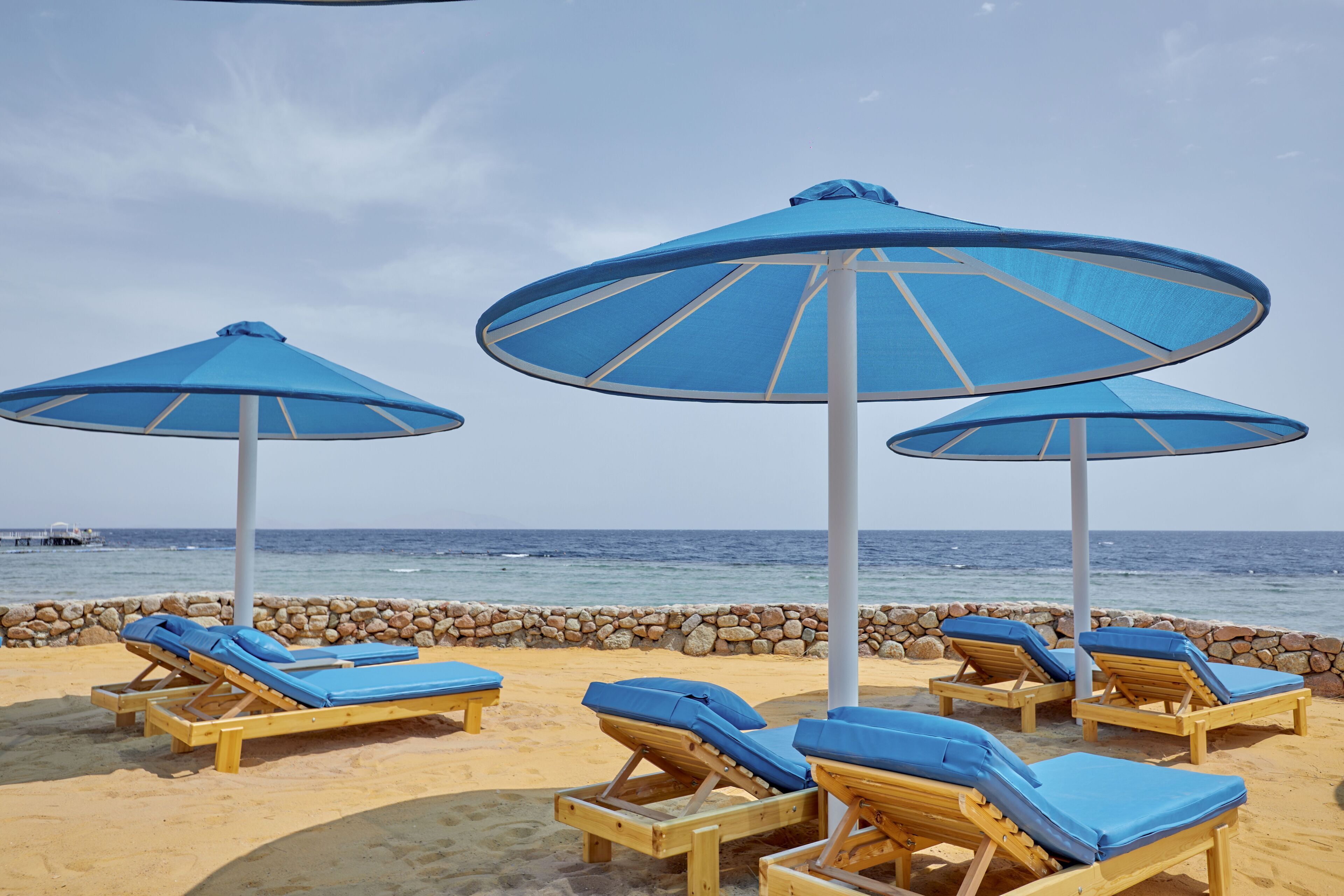 Un espace de détente aménagé sur la plage, avec des chaises longues en bois et des parasols bleus, offrant une vue sur l’océan.