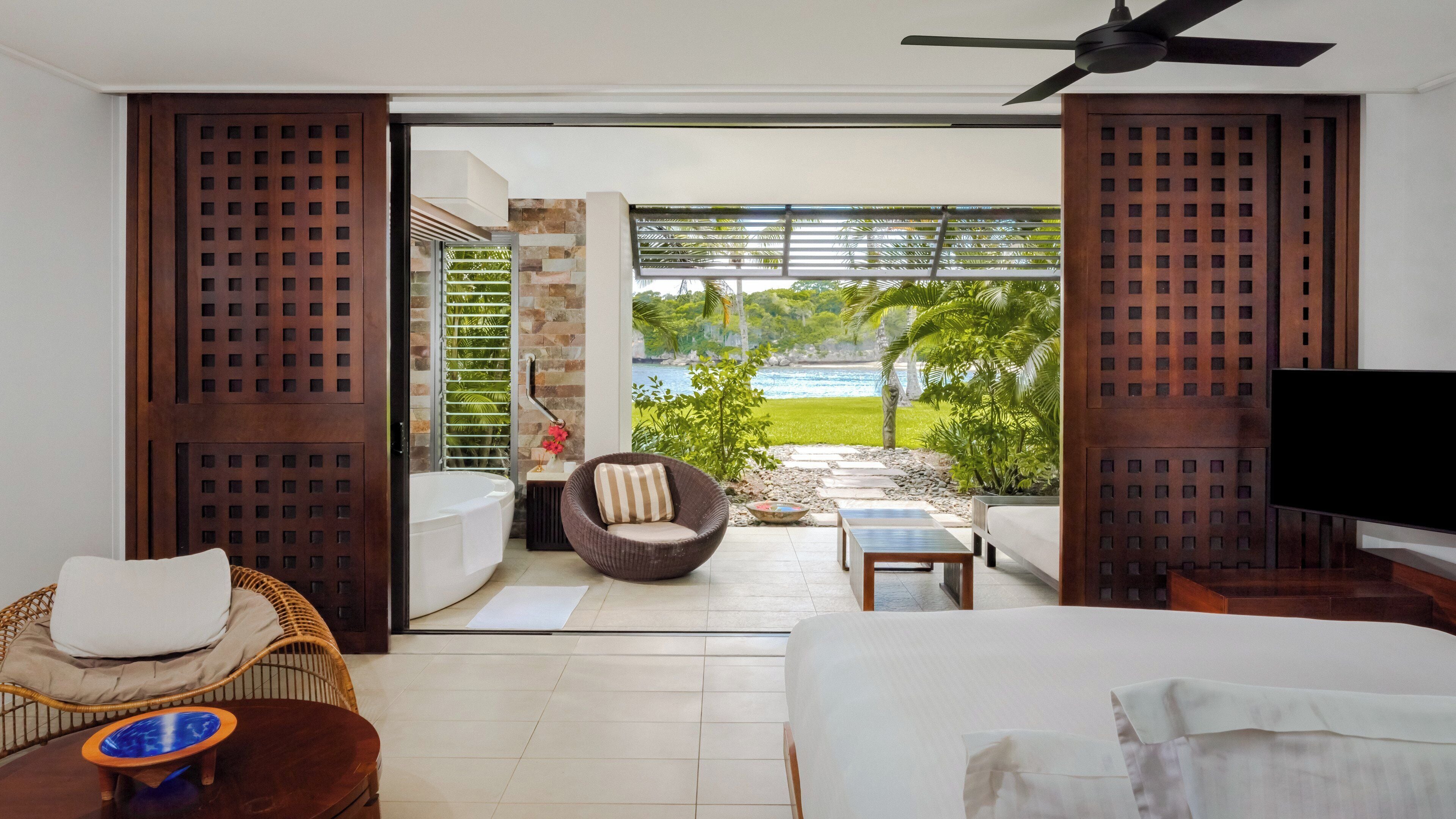 A hotel room with a bed, a TV, a wicker chair, a small table, and a view of a beach and palm trees through the open door.