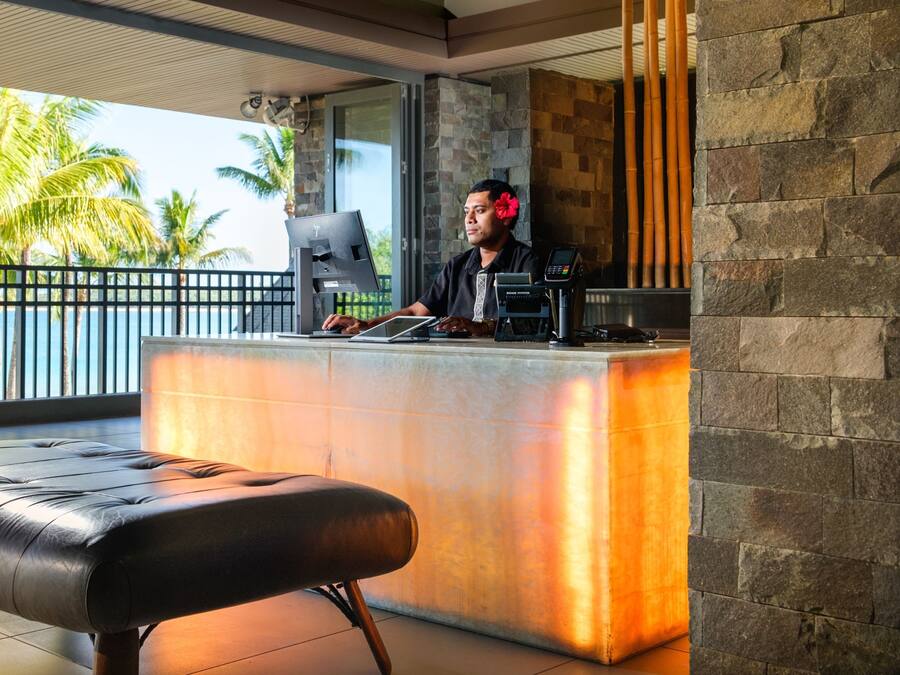 A reception area with a counter, a laptop, a cash register, and a barista. There is a bench in the foreground and a view of palm trees and a balcony outside.