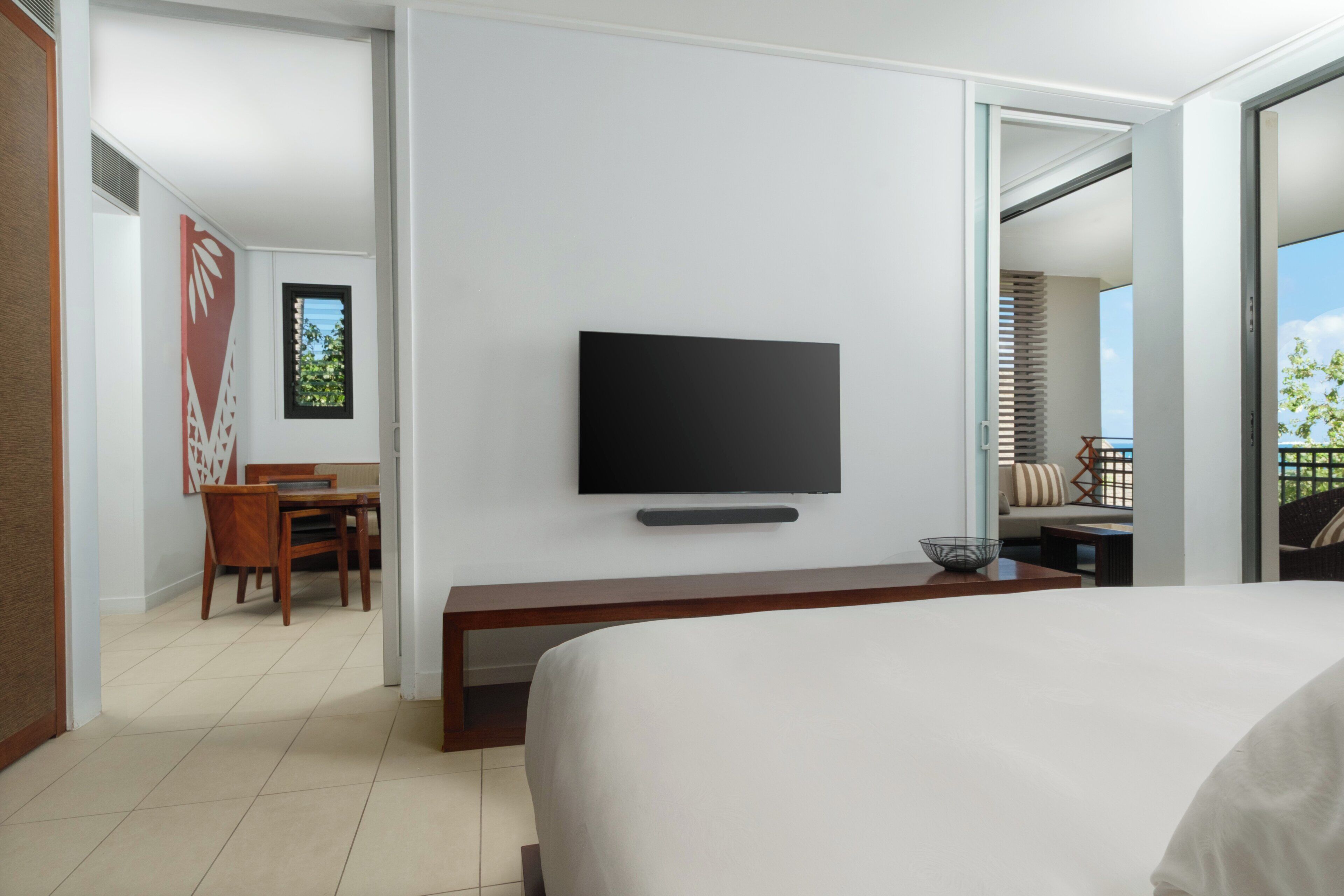 A hotel room with a bed, a TV mounted on the wall, a dining area with a table and chairs, and a balcony with a view.