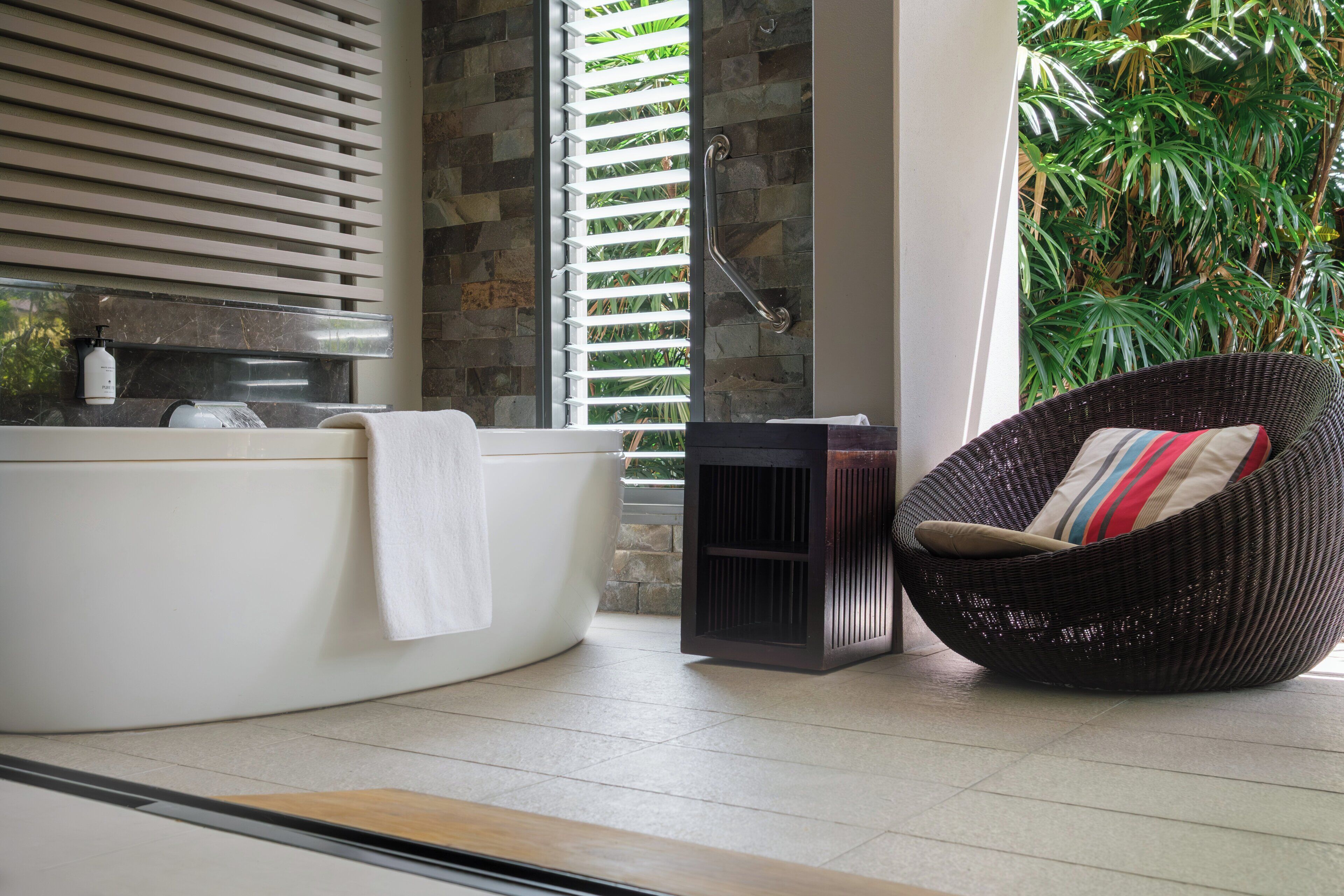 Un baño con bañera, una silla de mimbre con cojín y una ventana con persianas.