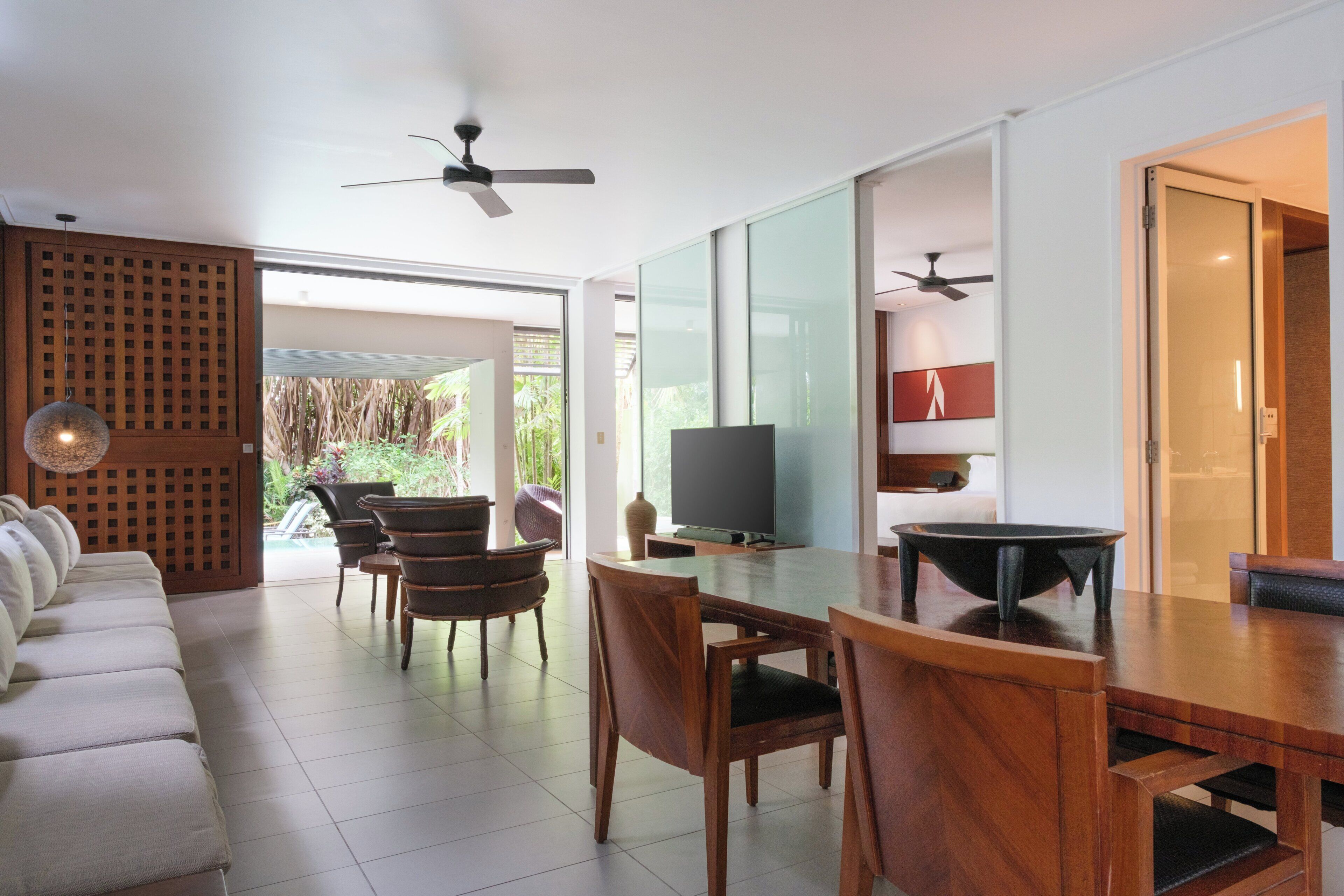 A modern living room with a sofa, dining table, chairs, and a TV. There is a sliding glass door leading to a balcony with plants.